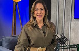 Mujer sentada en un sofá con camisa oliva, sonriente y un micrófono a su lado, en estudio con luz amarilla.