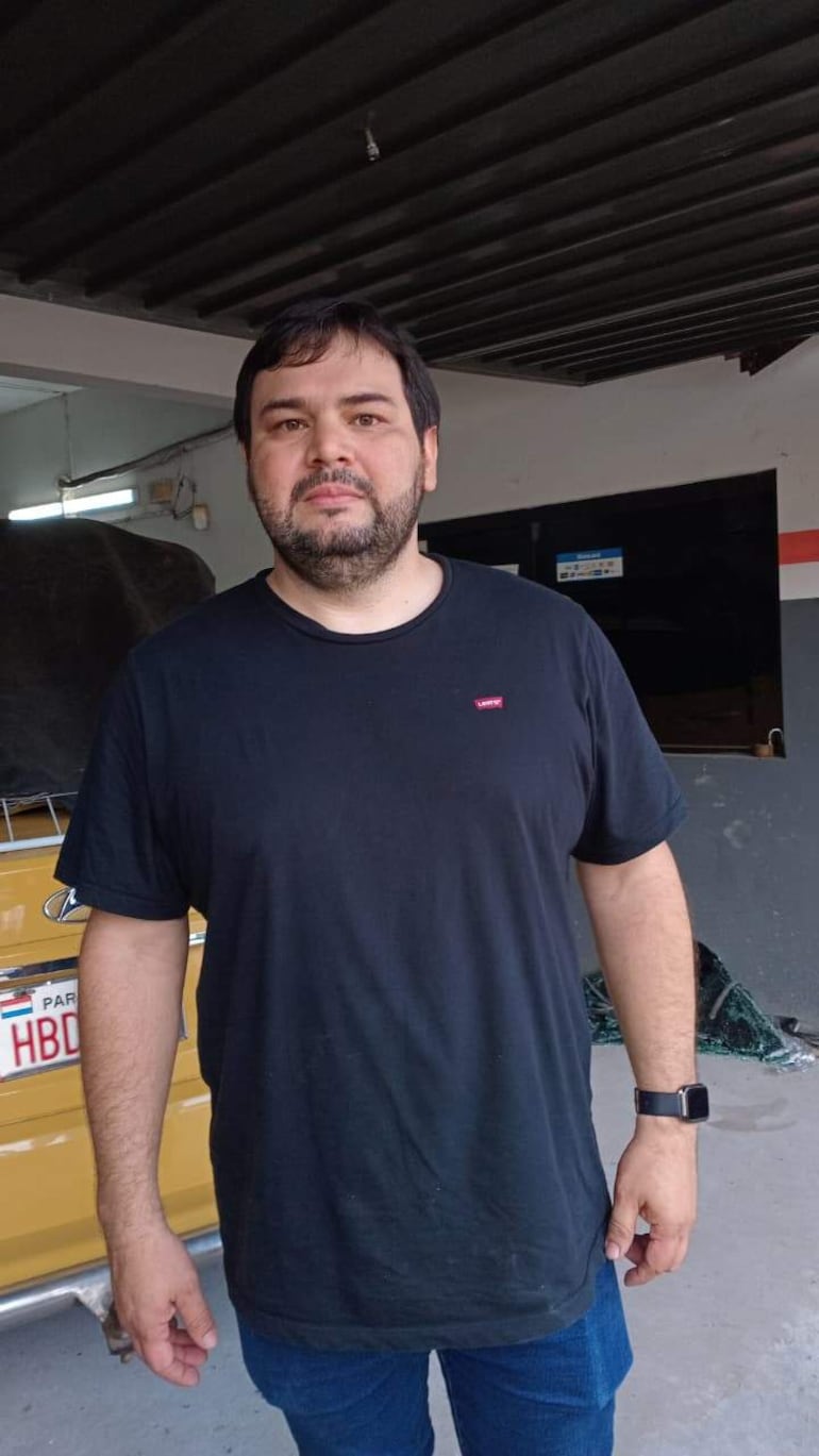 Hombre con camiseta negra y jeans de pie en un garaje, mirando hacia la cámara, mientras repara un parabrisas.