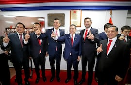 Baruja y cinco hombres en trajes oscuros con corbatas rojas levantan el dedo índice en un ambiente festivo decorado con la bandera paraguaya.