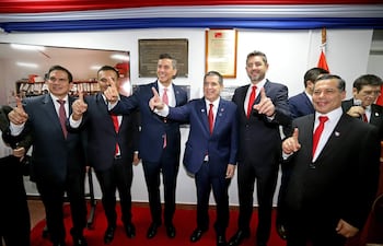 Baruja y cinco hombres en trajes oscuros con corbatas rojas levantan el dedo índice en un ambiente festivo decorado con la bandera paraguaya.