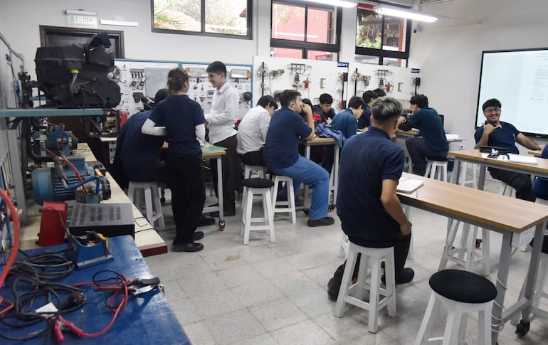 Joven con camisa blanca presenta en clase a estudiantes con camisetas azul marino en aula con equipo técnico.