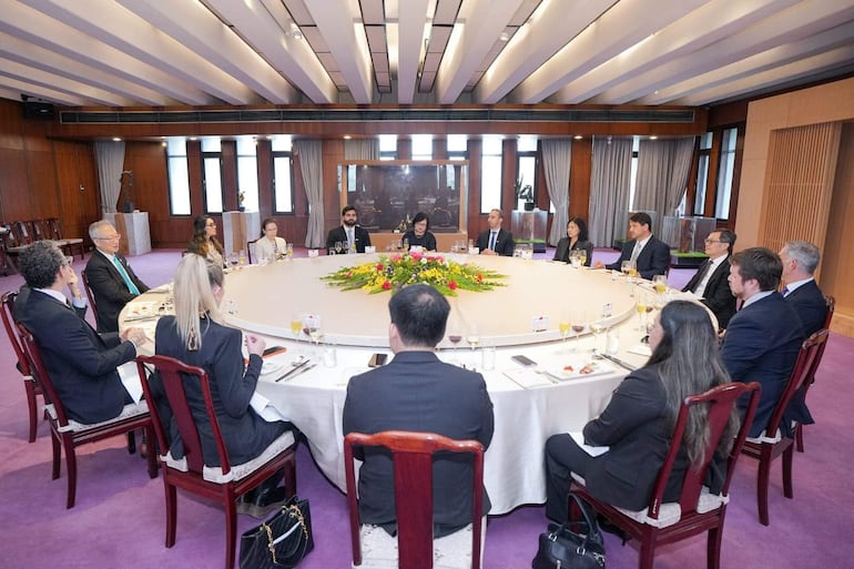 Reunión formal con 15 asistentes en trajes elegantes. Mesa circular decorada y banquete de comida. Conversación atenta entre los presentes.
