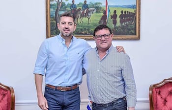 Hernán Ysidro Rivas con camiseta azul claro y Hercules Pedro Lorenzo Aliana Rodríguez en camisa de rayas, sonriendo en un despacho decorado con elementos patrios.