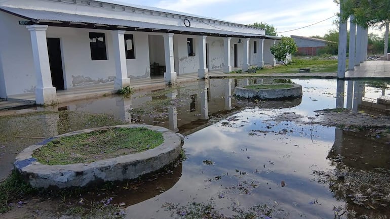 Area inundada frente a edificio escolar deteriorado, columnas blancas y ventanas abiertas, con espacio verde al fondo.