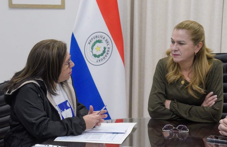 María Teresa Barán, vestida con blusa verde, escucha atentamente a la presidenta de la APE, quien usa chaqueta oscura y sostiene un bolígrafo en una oficina.