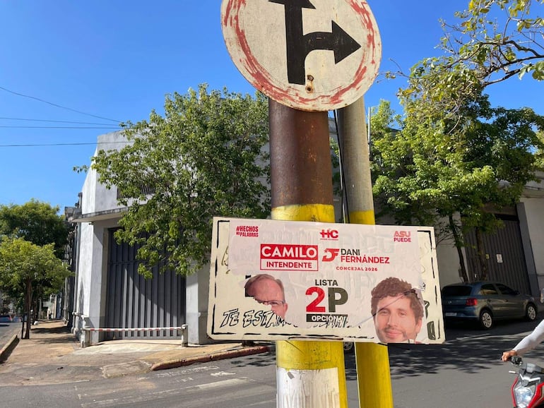Cartel electoral en poste con rostros de Camilo Intendente y Dani Fernández, fondo urbano despejado y árboles.