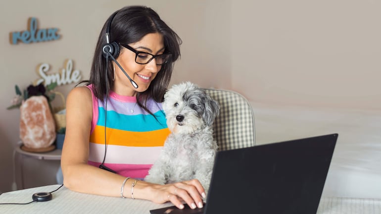 Mujer con camiseta multicolor, usando auriculares, sentada ante un escritorio con un perro blanco y gris a su lado.