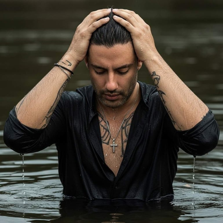 Hombre con cabello recogido y camisa negra húmeda, agachado con las manos en la cabeza, en un entorno natural acuático.