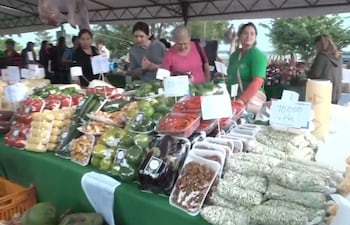 Tres mujeres en el mercado al aire libre. Una con camiseta verde vende mientras las otras examinan productos frescos en mesas.