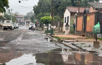 Dos personas conversan en la calle, mientras un vehículo estacionado muestra agua acumulada. Un árbol y un recipiente de basura son visibles.