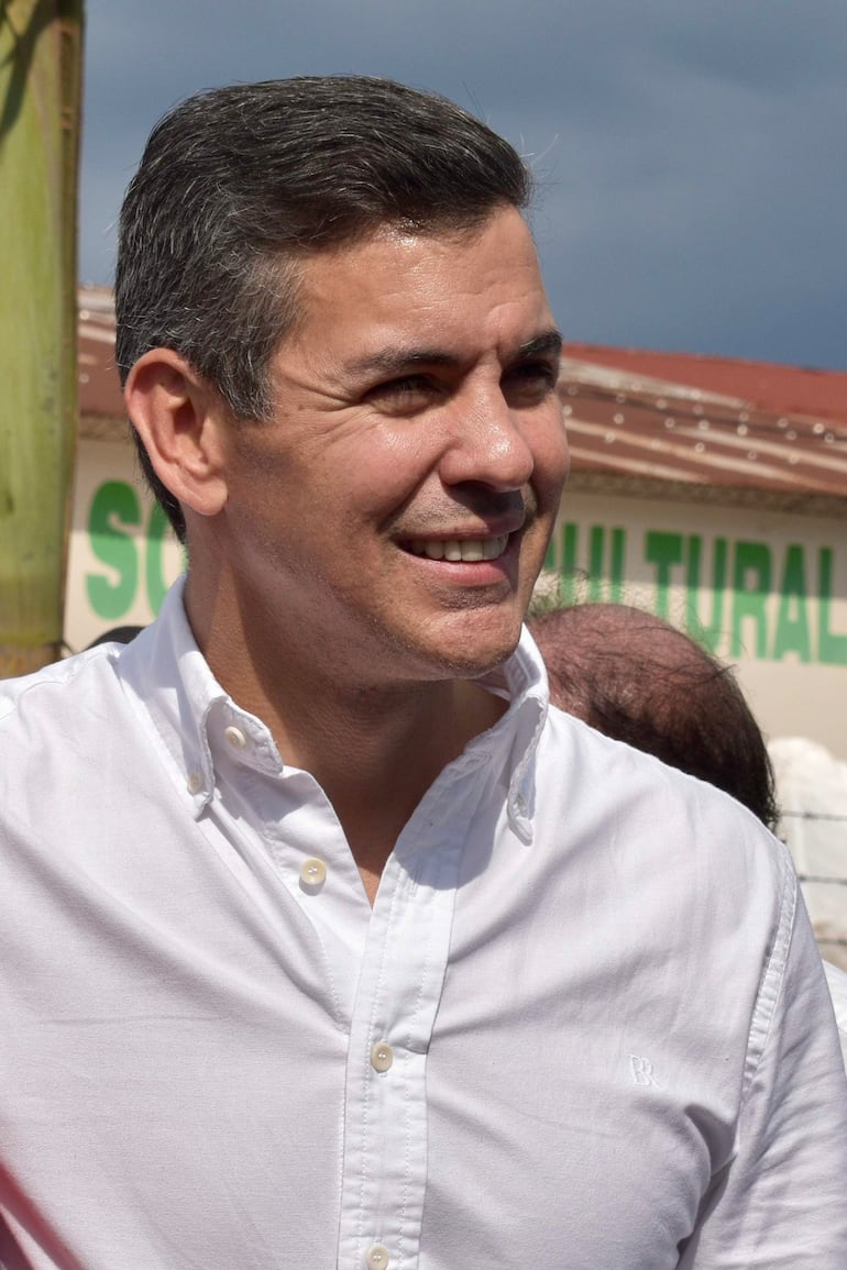 Santiago Peña sonriente con camisa blanca, interactuando en evento al aire libre, rodeado de personas en un ambiente social.