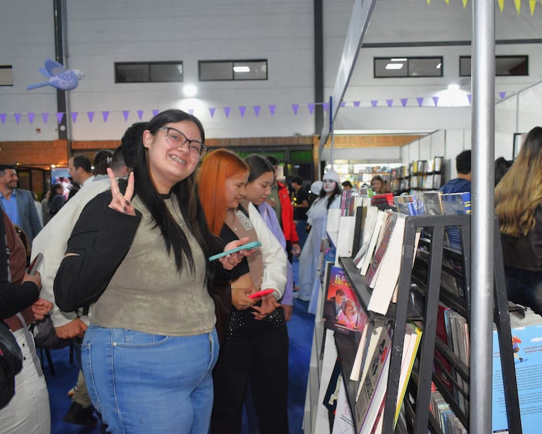 Mujer con gafas sonriente haciendo un gesto de paz, rodeada de personas en un ambiente decorado con banderines morados.