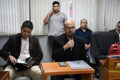 Hombre con saco negro habla al micrófono, rodeado de otros hombres en ambiente de reunión, con documentos en mano.