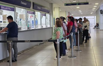 Fila de pacientes en ropa casual, esperando en la farmacia del hospital. Una persona atiende desde el mostrador.