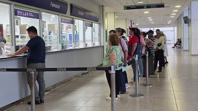 Fila de pacientes en ropa casual, esperando en la farmacia del hospital. Una persona atiende desde el mostrador.