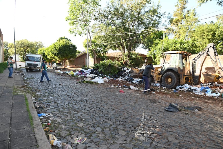 Dos hombres limpian basura en la calle adoquinada. Un tercero toma notas cerca de un camión de basura.