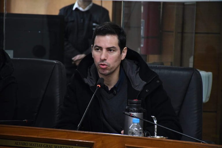 Álvaro Grau, hombre de cabello oscuro en abrigo negro, habla con micrófono en ambiente de deliberación, acompañado de un asistente.
