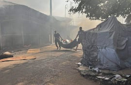 Dos hombres con ropa oscura cargan un bulto mientras el humo y las llamas envuelven un mercado en Itauguá.