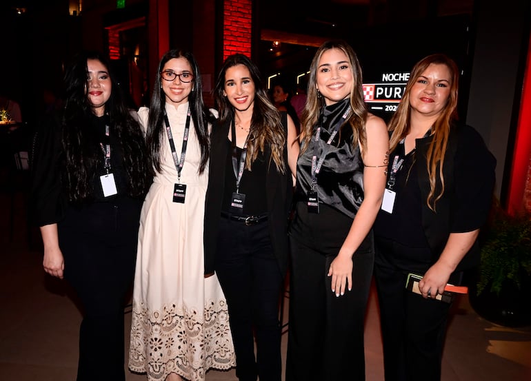 Cinco mujeres sonrientes en atuendo negro y blanco, disfrutan de un evento iluminado con luces cálidas en ambiente festivo.