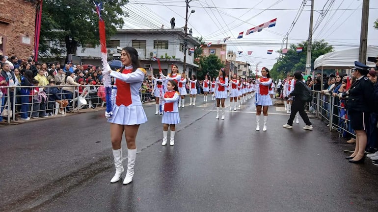 Grupo de jóvenes y una niña con trajes blancos y rojos marchando en un desfile, sosteniendo objetos decorativos bajo un clima nublado.