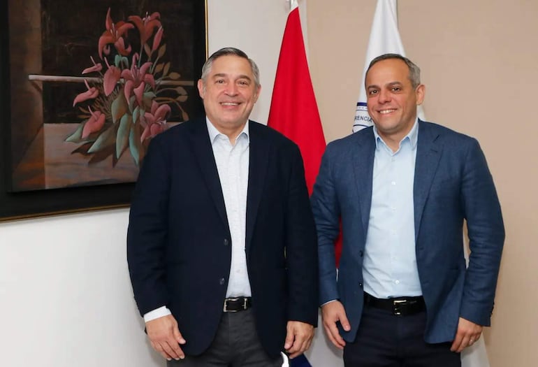 Luis Ramírez y Camilo Benítez sonríen en el despacho del MEC, vestidos con blazers oscuros, rodeados de banderas.