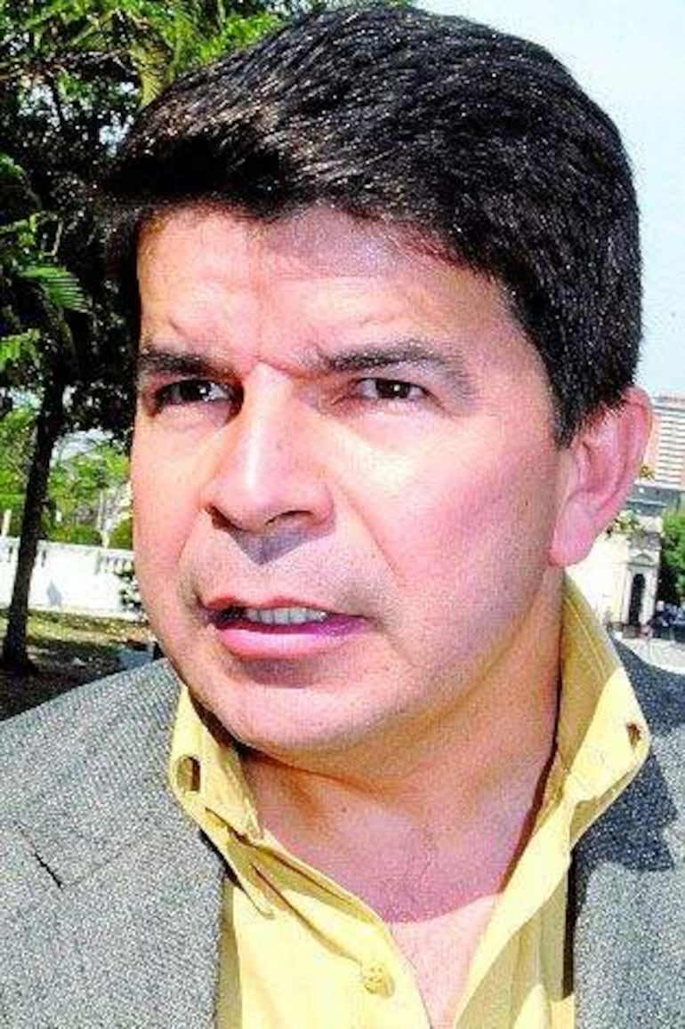 Hermenegildo Cohene con camisa amarilla y saco gris, en conversación intensa entre verdes plantas al fondo.