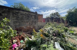 Ambiente descuidado con montículos de tierra y basura, muros de ladrillo en el fondo y una cruz visible entre edificios.