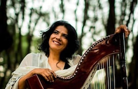 Norma Ortega sonríe mientras sostiene un arpa en un parque, con árboles de fondo y vestimenta clara y adornada.