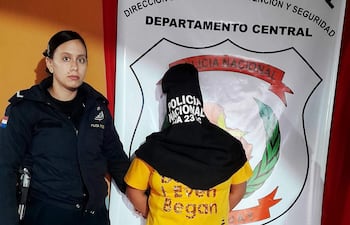Policía sostiene a un individuo con pasamontañas negro y camiseta amarilla, mientras una oficial supervisa la situación en un ambiente claro.