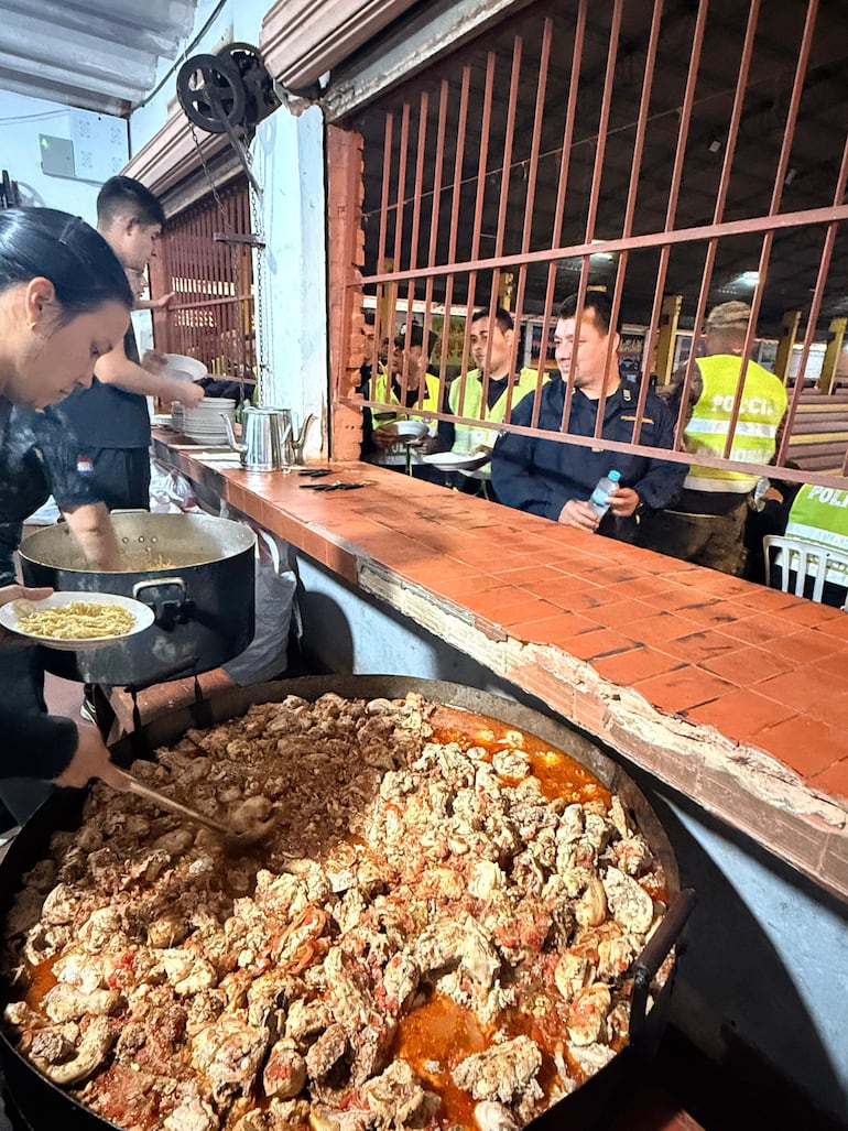 Mujer con cabello atado cocina en olla grande, mientras hombres en chalecos policiales observan desde atrás, esperando comida.