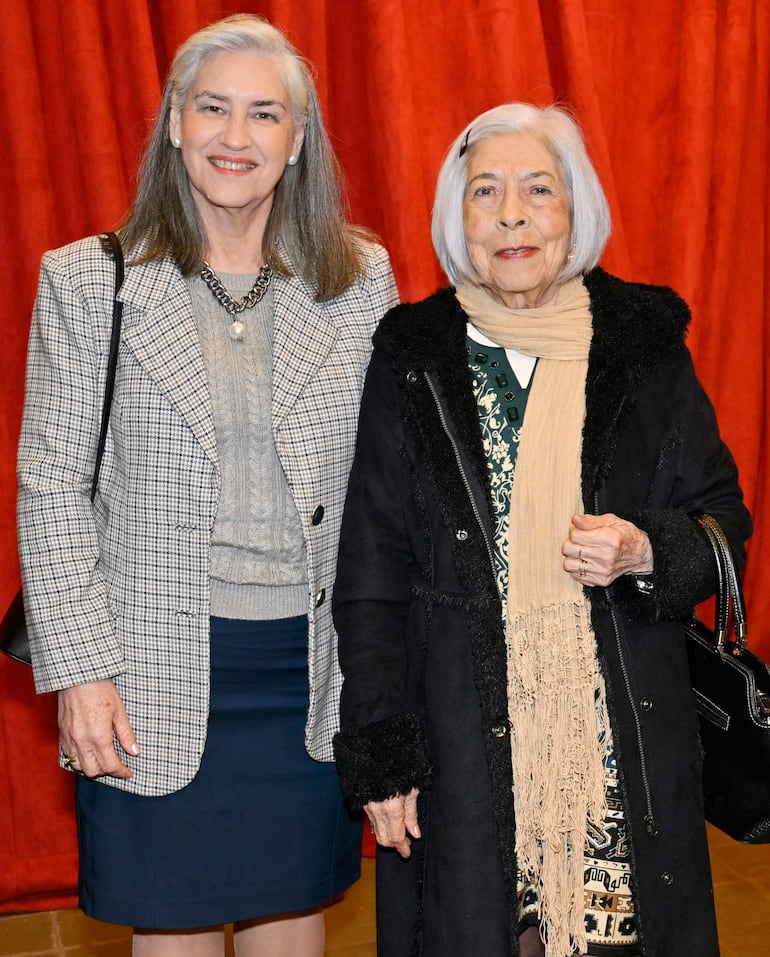 Dos mujeres sonriendo frente a una cortina roja, la de la izquierda con abrigo de cuadros y cabello gris, y la otra con abrigo oscuro y bufanda clara.