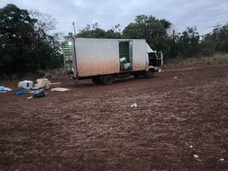 Camión blanco con puerta trasera abierta en terreno seco, rodeado de bolsas plásticas y cartones, sin personas visibles.