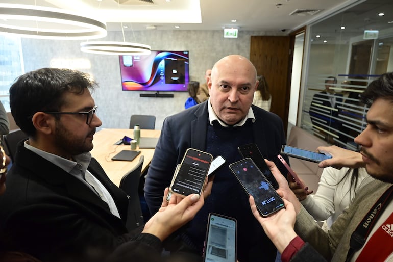 Hombre corpulento con chaqueta oscura y camisa blanca rodeado de periodistas con teléfonos y cámara en un ambiente moderno.
