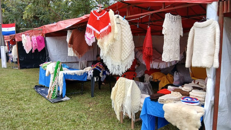 Feria al aire libre con toldos coloridos, mesas exhibiendo productos de lana como bufandas y ponchos en un entorno natural.