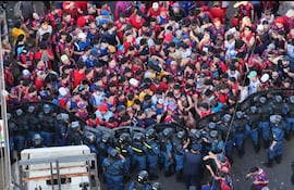 Multitud de aficionados con camisetas rojas y azules, gesticulando cerca de policías antimotines en ambiente urbano.