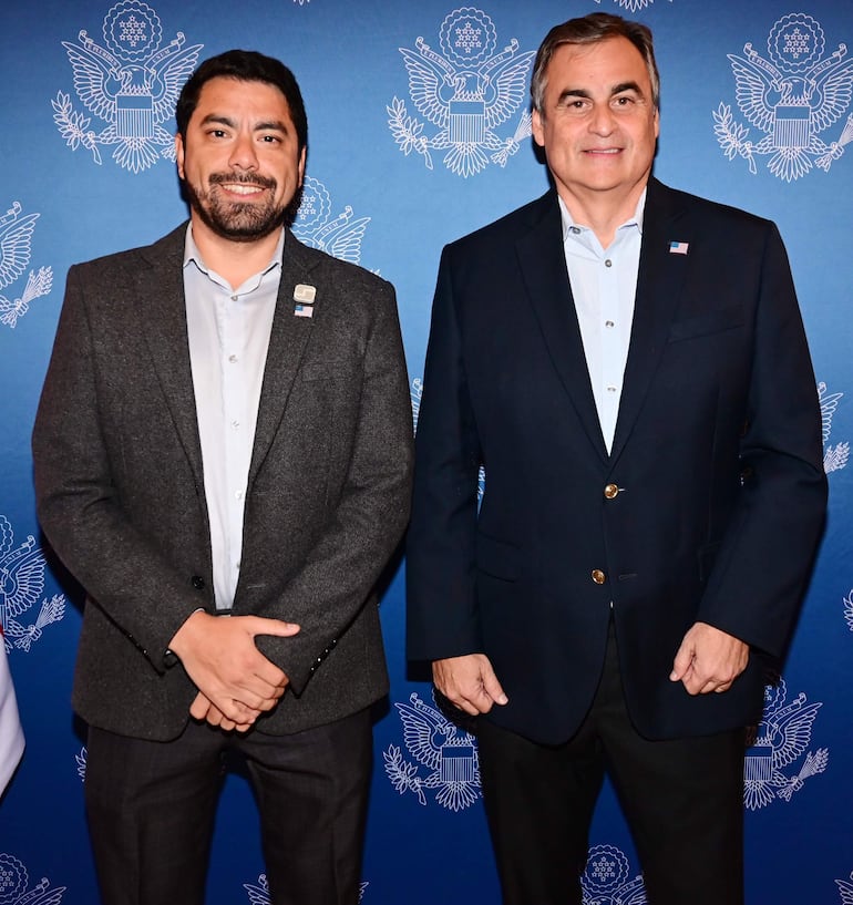 Hombres sonrientes en blazers frente a fondo azul con el emblema del gobierno de EE. UU.