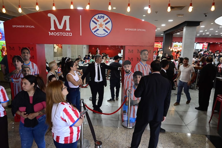 Julio Enciso, entre otros, en ambiente moderno de La Joya Mostaza, con expresiones felices y ropa de hinchas de fútbol.