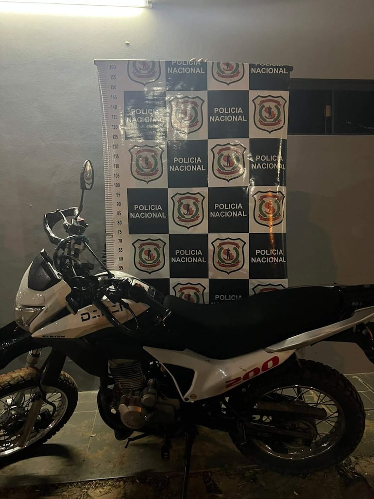Moto blanca y negra con banner de la Policía Nacional detrás, en un ambiente exterior iluminado artificialmente.