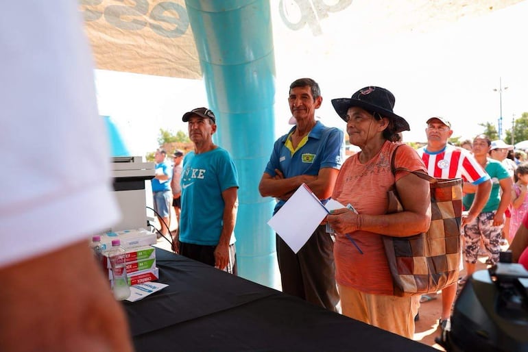 Grupo de personas en evento al aire libre, mujer mayor con sombrero y cuaderno escucha atentamente mientras un hombre habla.