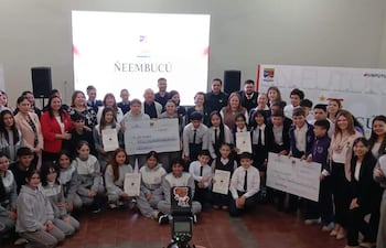 Grupo diverso de estudiantes sostiene cheques y diplomas, con expresión de alegría en un evento de premiación formal.