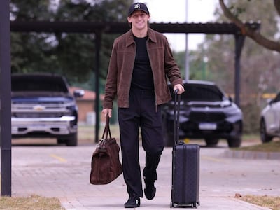 Hombre con chaqueta marrón, camiseta negra y pantalones oscuros, camina hacia la cámara, sosteniendo un maletín y maleta en un entorno urbano.