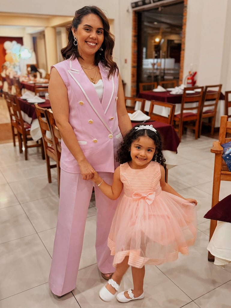 Mujer con vestido rosa claro toma de la mano a niña en vestido rosa con cinta, ambas sonrientes en un ambiente festivo.