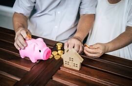 Pareja de adultos mayores ahorrando monedas en una alcancía junto a una maqueta de casa, representando el sistema de pensiones y jubilaciones en Paraguay.