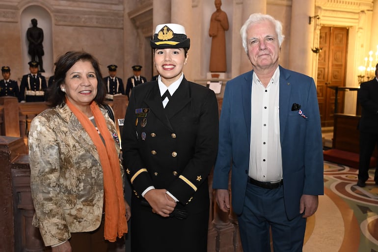 Mujer con chaqueta clara y bufanda naranja sonríe junto a oficial naval y hombre con traje azul en evento decorado con estatuas.