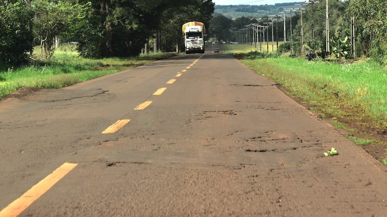 Camión circulando por carretera con baches y desgaste, rodeado de vegetación y árboles en un entorno rural.