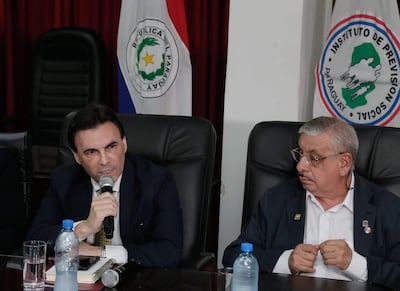 Carlos Pereira y Isaías Fretes en conferencia, el primero con micrófono y traje oscuro, mientras el segundo escucha atentamente con traje claro.