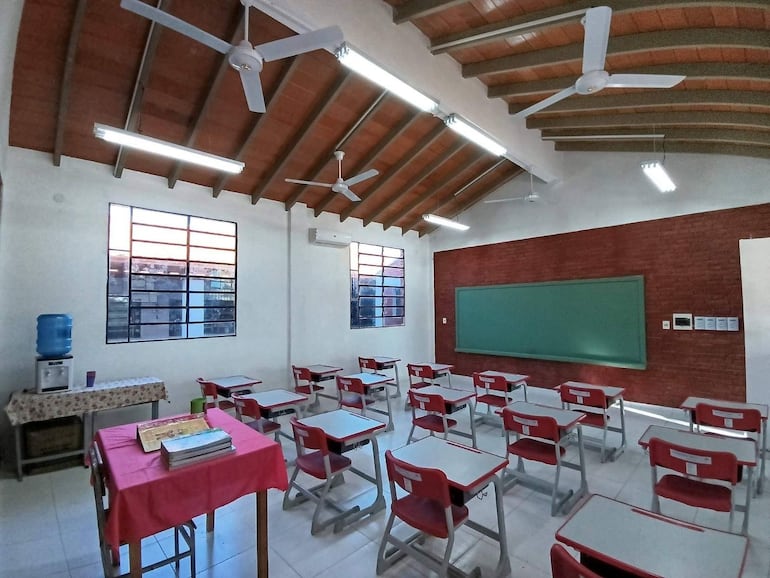 Aula moderna y luminosa con escritorios y sillas rojas alineadas, dispensador de agua azul a la izquierda, ambiente interior. Sin personas.