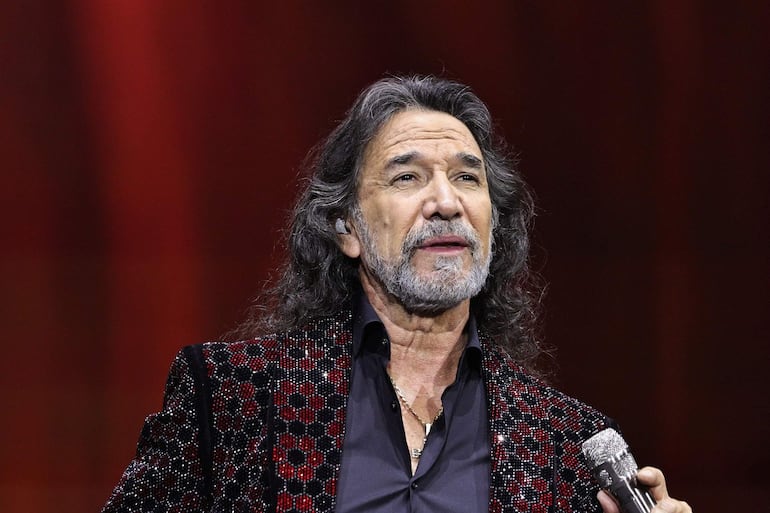 Marco Antonio Solís, con cabello largo y chaqueta brillante, habla al público sosteniendo un micrófono en un escenario iluminado.