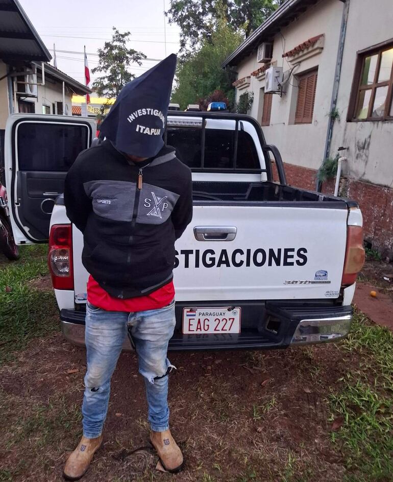 Hombre con capuchón negro de 'INVESTIGACIONES ITAPÚA' y chaqueta de colores oscuros, frente a una camioneta policial en un entorno verde.
