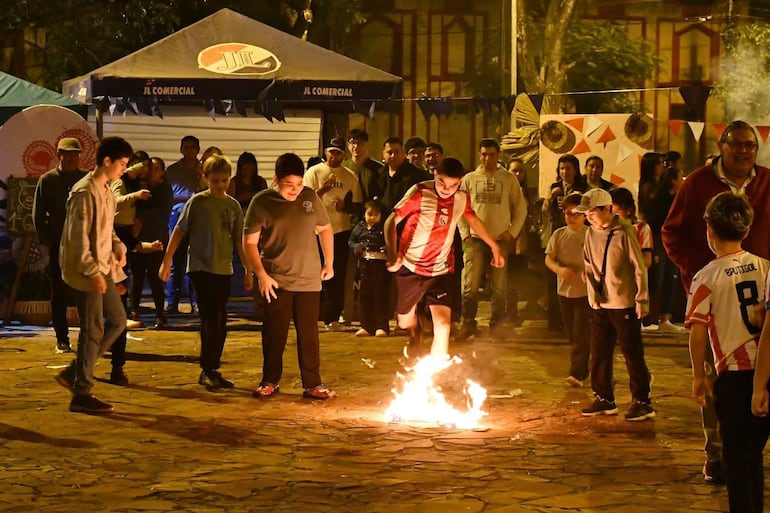 Niños saltan sobre fuego, uno lleva camiseta roja y blanca del Club Olimpia, ambiente festivo y de celebración.
